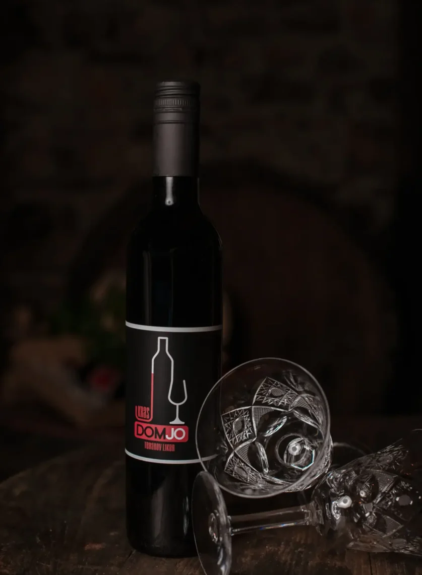 Elegantna temna produktna fotografija steklenice vina DOMJO ob kristalnem kozarcu.