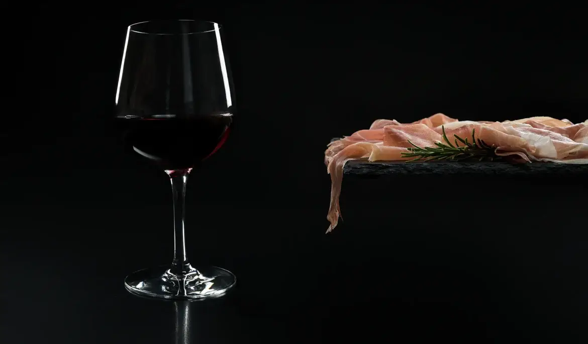 Kozarec rdečega vina terana ob narezku kraškega pršuta z rožmarinom na črni skrilavčevi plošči.
