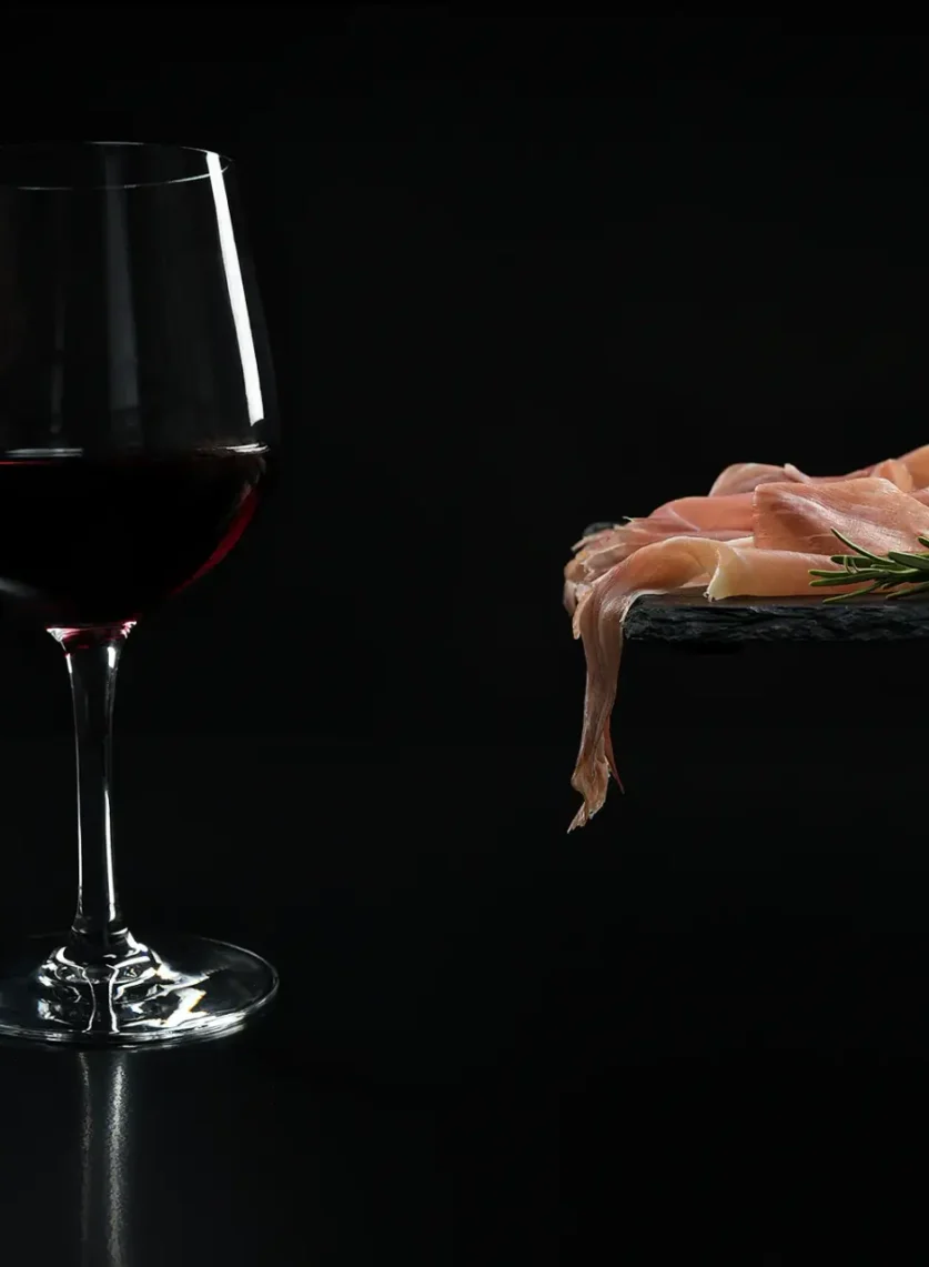 Kozarec rdečega vina terana ob narezku kraškega pršuta z rožmarinom na črni skrilavčevi plošči.