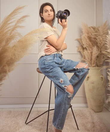 fotografinja-vani-foto-studio-kreativa (1) Fotografinja Vani sedi na visokem stolu v svojem studiu s fotoaparatom v roki, obdana z estetsko dekoracijo iz pampaške trave.Fotografinja Sežana
